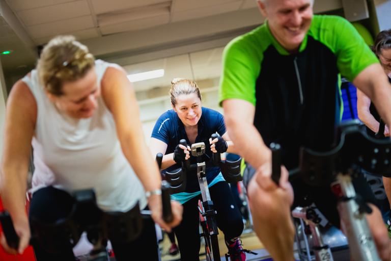 Image shows a small group of three people on studio cycling bikes taking part in an exercise class. 
