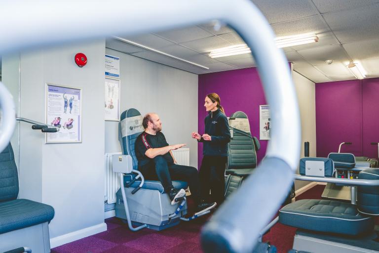 Image shows man in the distance sitting on toning chair, being instructed by a toning instructor on how to use the machine. 