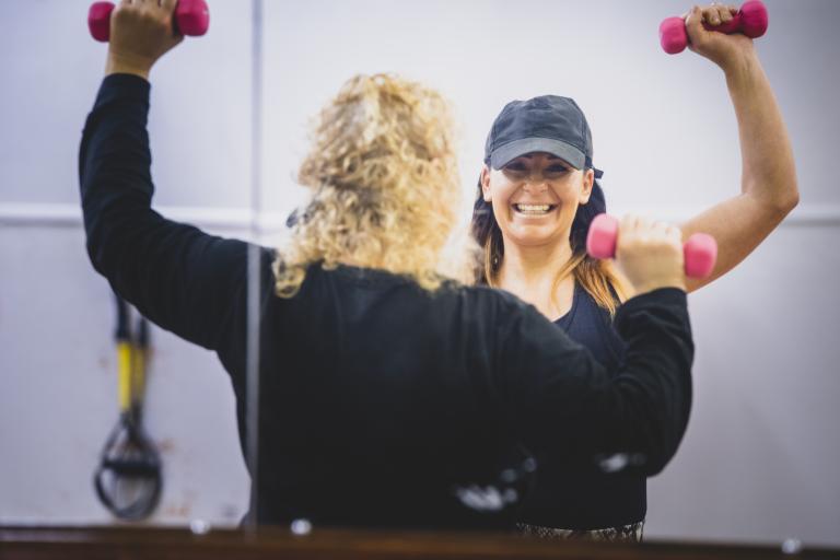 Image shows mirror image of instructor with woman lifting dumbells as part of an exercise class.