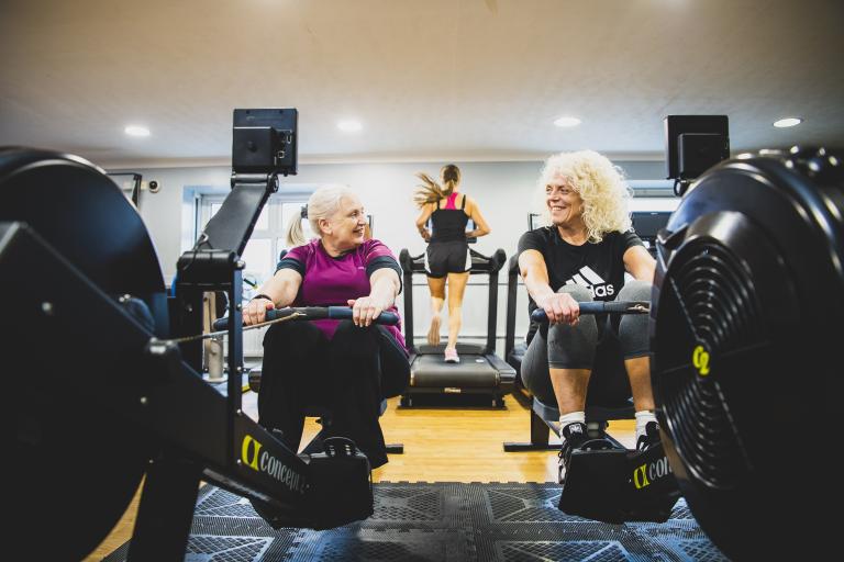 Two women chatting, sat next to each other on rowing machines.
