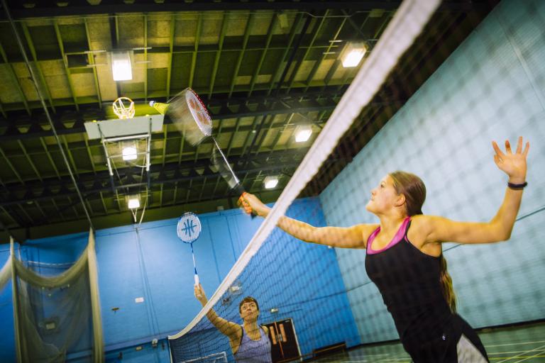 Image shows girl holding badminton racket, hitting shuttlecock over net in sportshall. 