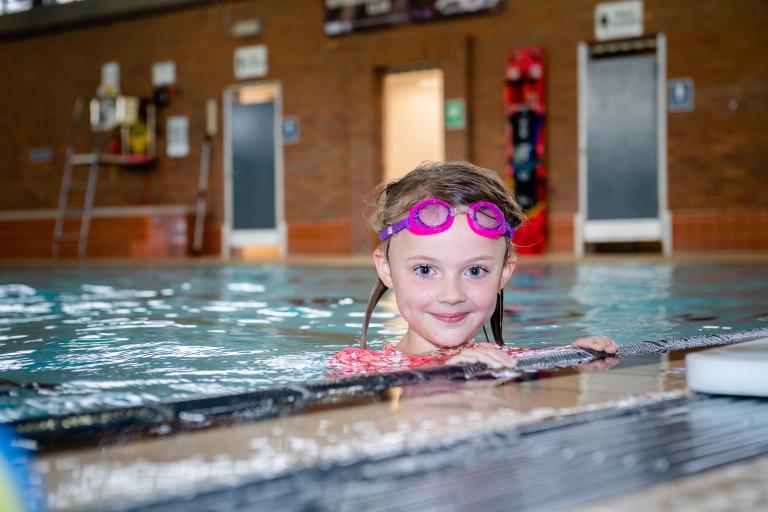 Image shows girl in swimming pool, holding onto poolside with goggles on. 