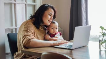 A woman uses her computer while holding a young child