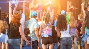 A crowd of people, including adults and children, are enjoying a live outdoor concert, raising their hands towards the stage in excitement.