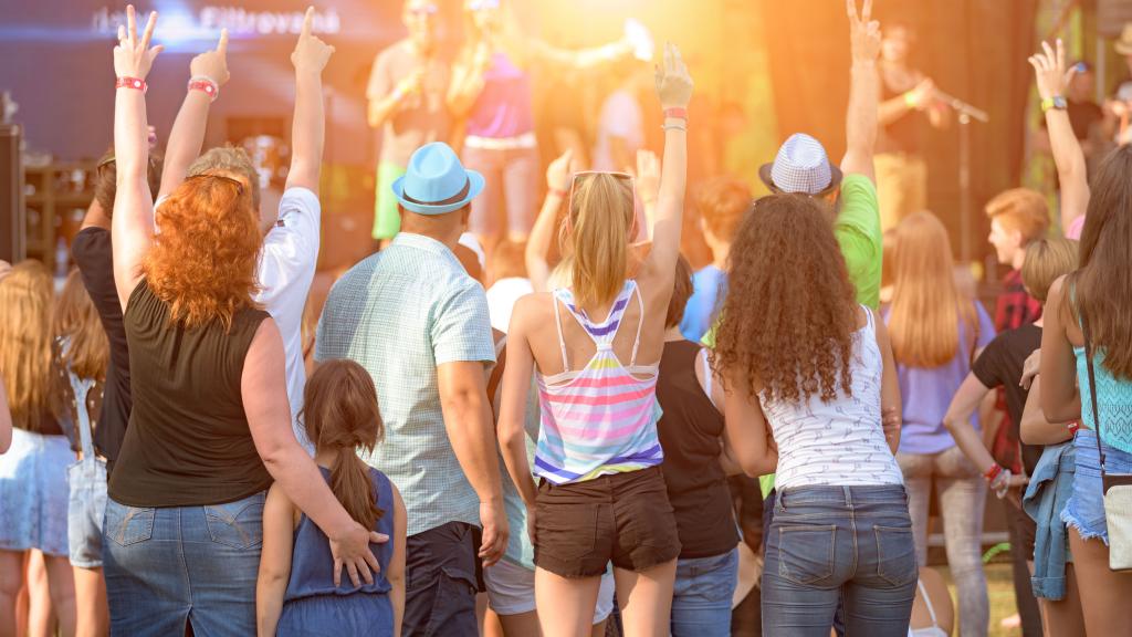 A crowd of people, including adults and children, are enjoying a live outdoor concert, raising their hands towards the stage in excitement.