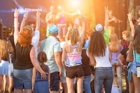 A crowd of people, including adults and children, are enjoying a live outdoor concert, raising their hands towards the stage in excitement.